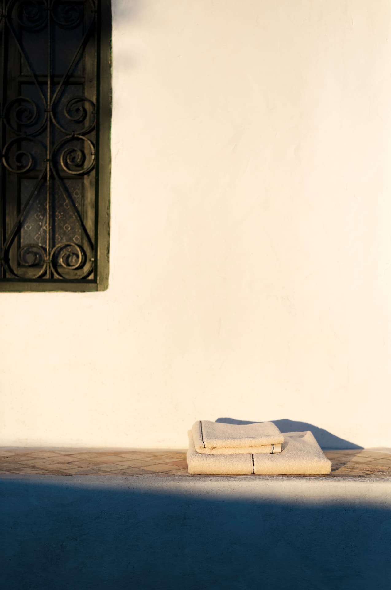 Folded towels on a tiled ledge against a white wall with an ironwork window