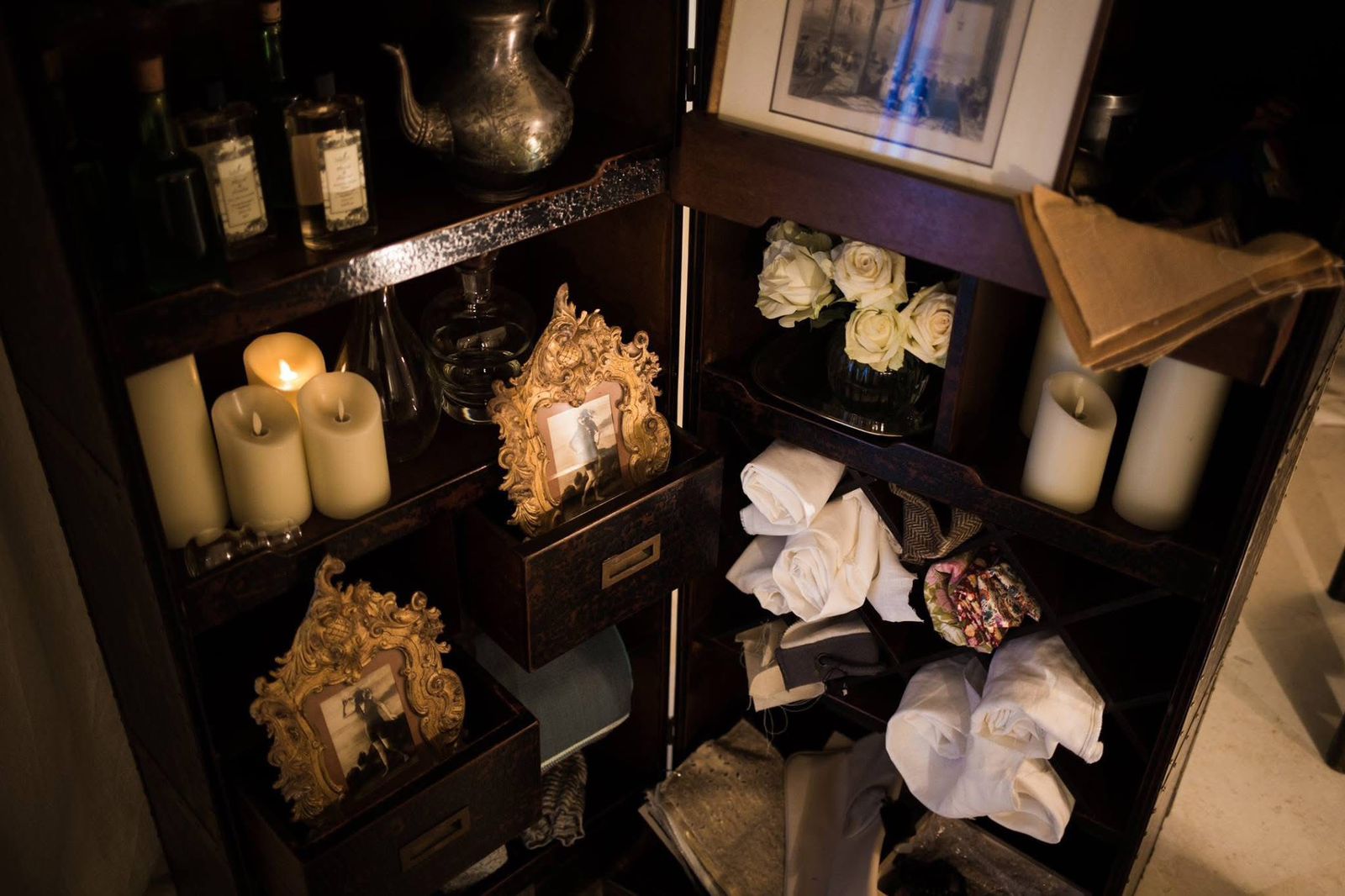 Cabinet arrangement with candles, framed photographs, flowers and folded textiles
