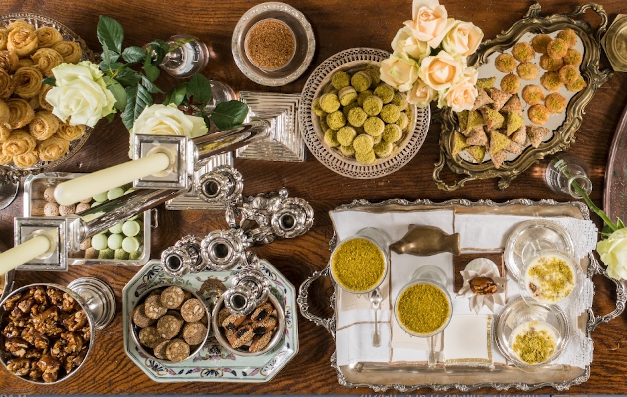 Overhead table of patisserie with silver trays of pistachio sweets, pastries, roses and candles on a wooden surface