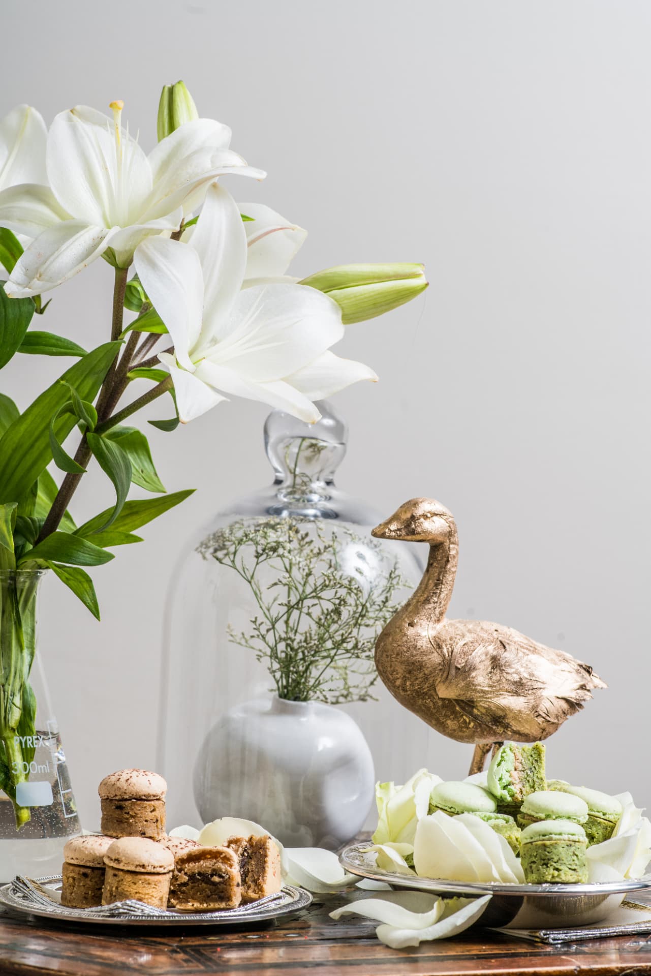 Still life with white lilies, a gilt bird sculpture, a glass cloche and plates of pistachio sweets
