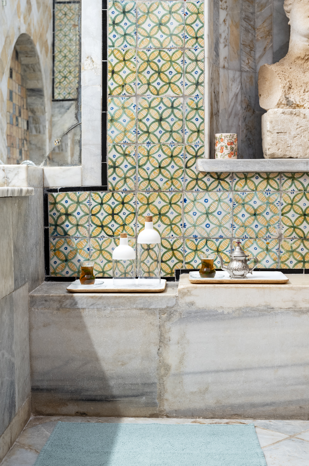 Patterned tiles with tea glasses, a silver teapot and white bottles on marble