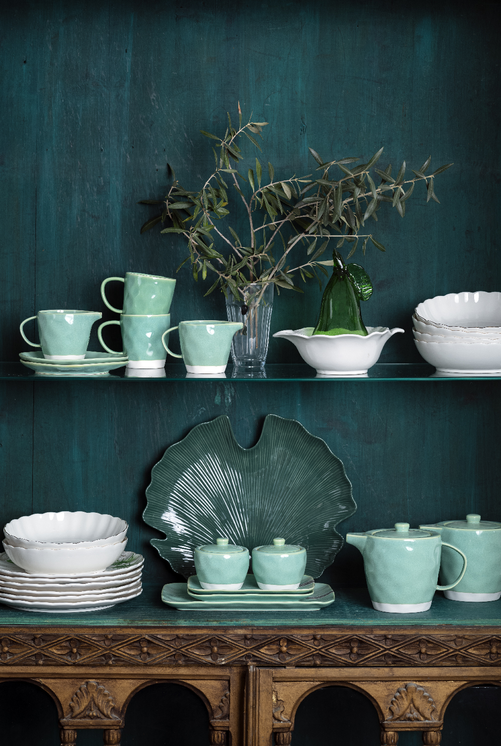 Celadon ceramics and white bowls on glass shelves against a dark teal wall